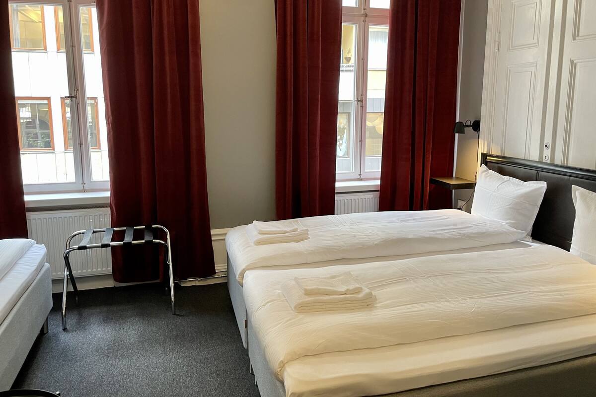 Habitación de hotel con dos camas, un radiador, una ventana con cortinas y una mesita de noche con lámpara.