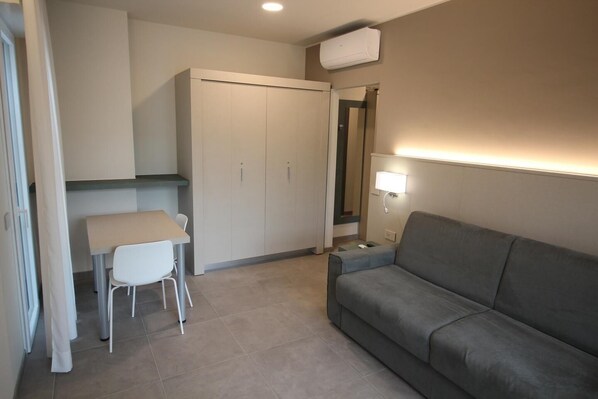 A compact room with a grey sofa, a small table with two chairs, a wardrobe, and a wall-mounted lamp.