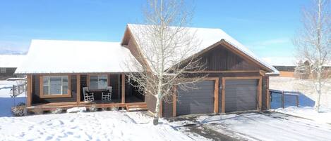 A wooden cabin with a covered porch, a garage, and a snow-covered landscape.
