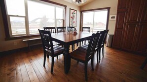 Une salle à manger avec une table et des chaises en bois, de grandes fenêtres et un lustre.