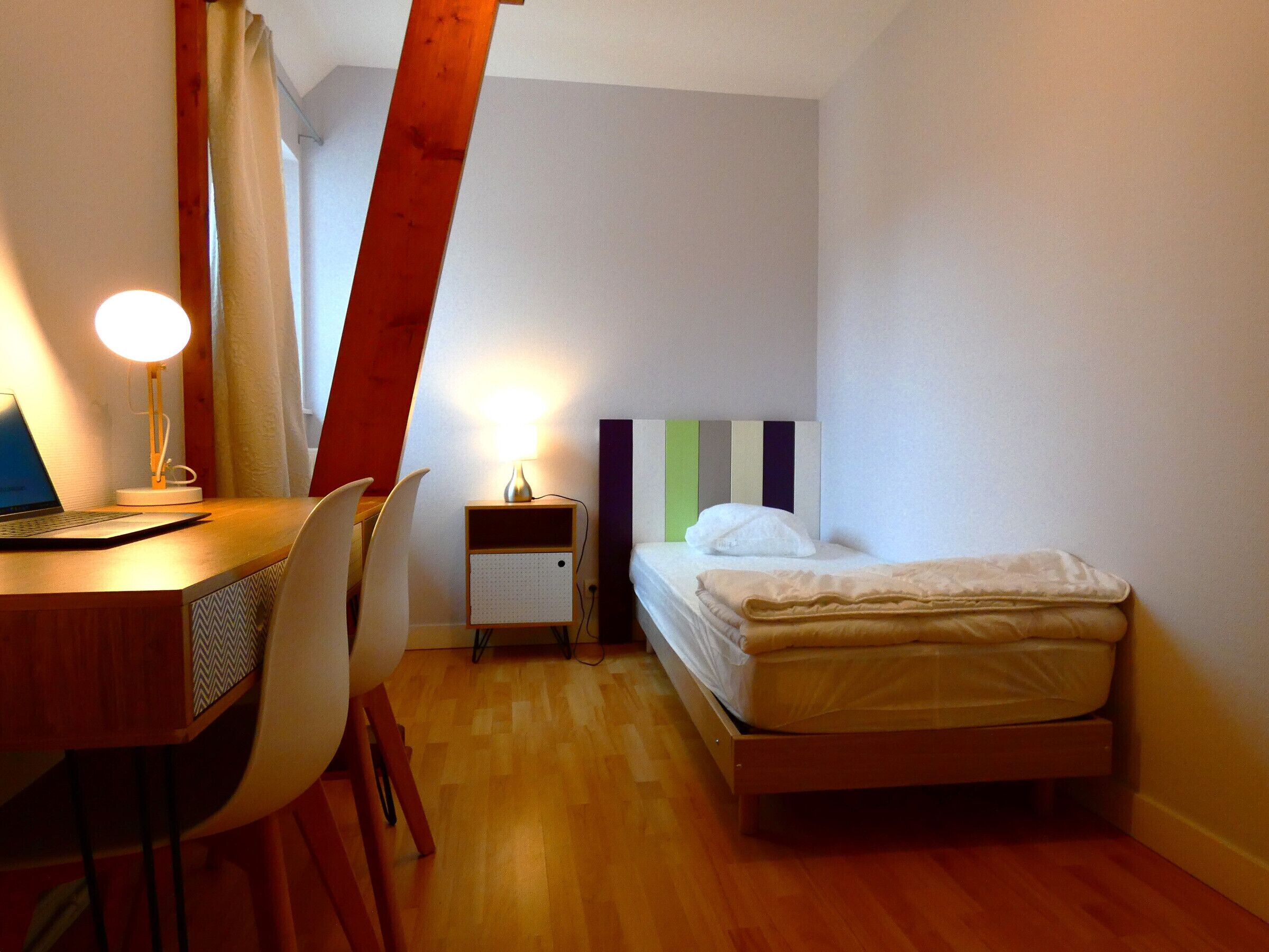 Una camera da letto piccola e ordinata, con una scrivania in legno, una sedia, un letto con testiera a righe e un comodino con una lampada.
