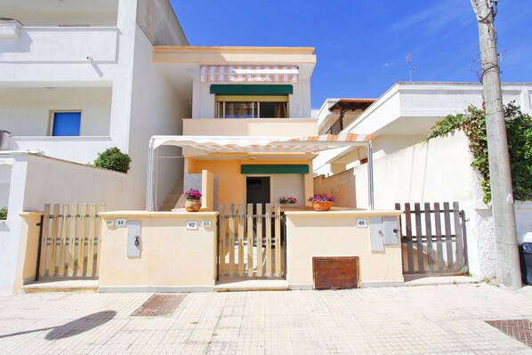 A modern two-story building with a gated entrance, a covered porch, and a small balcony with potted plants.