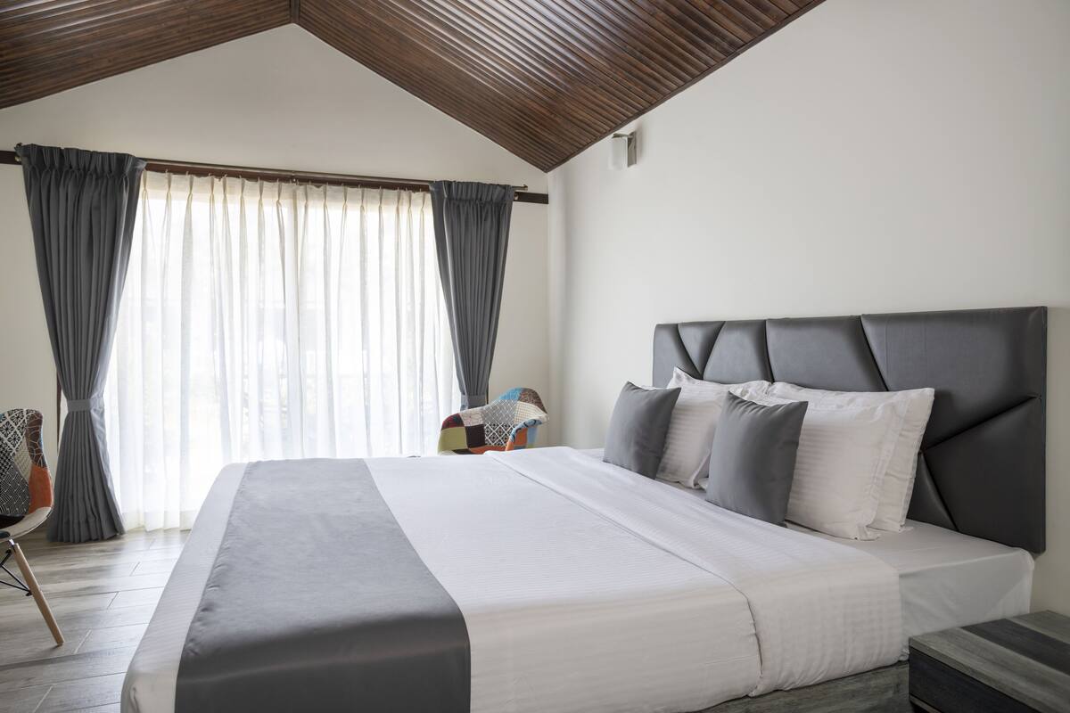 Quarto moderno com uma cama grande, teto de madeira e vista para o exterior através de cortinas leves.