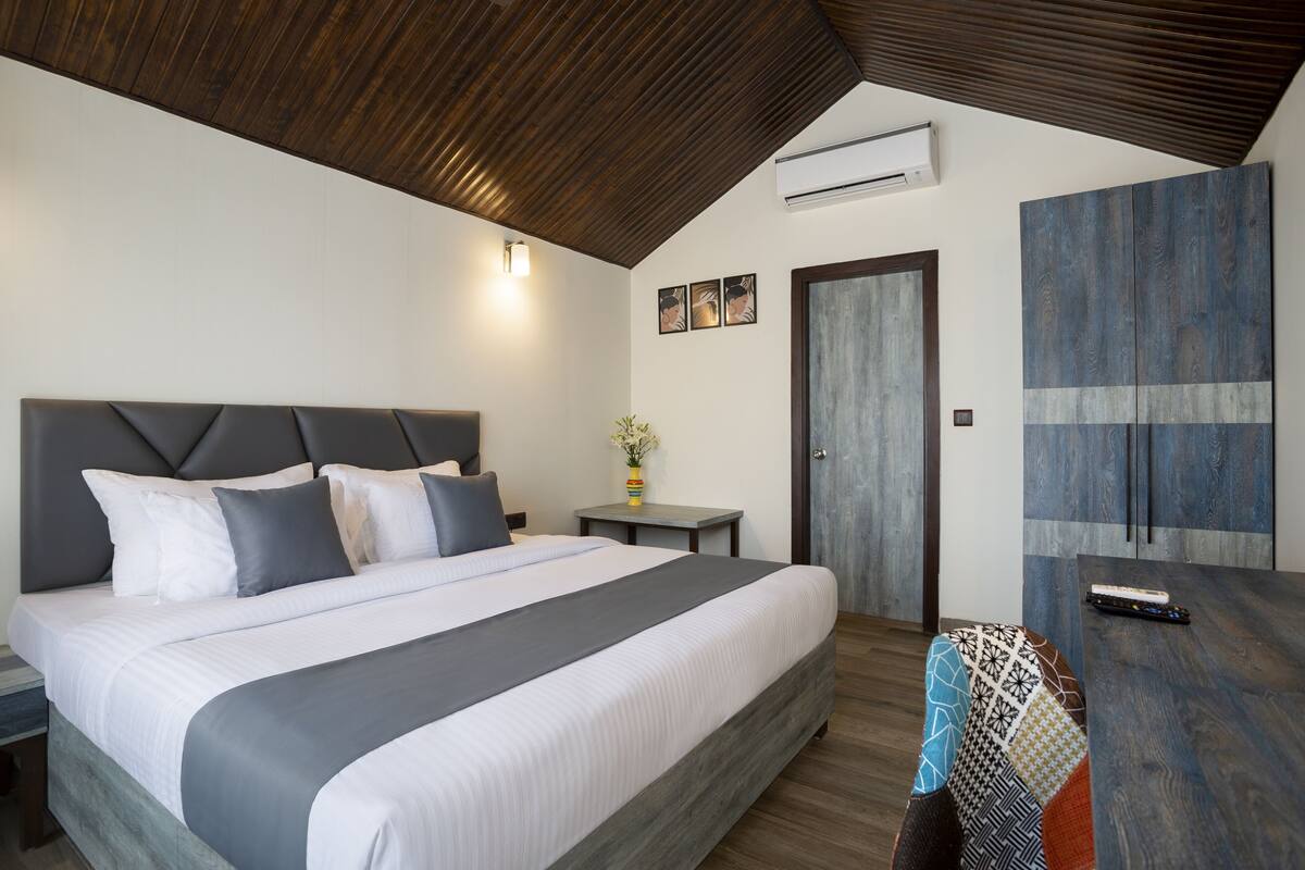 Quarto moderno com uma cama grande, teto de madeira, uma mesa pequena com um vaso, e uma porta.