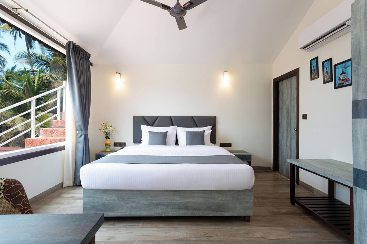 Quarto moderno com cama grande, ventilador de teto, varanda com guarda-corpo e vista para palmeiras.