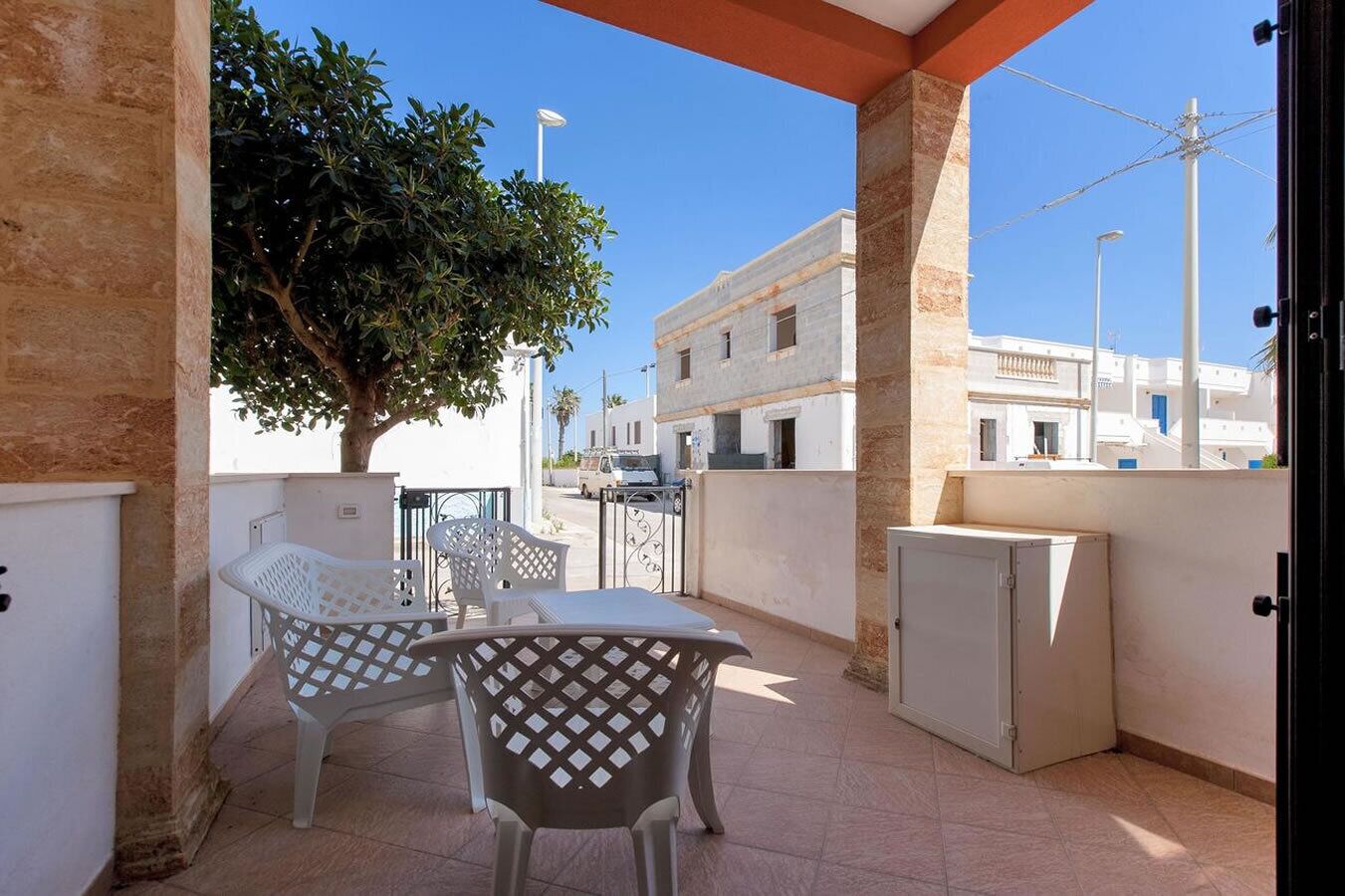 Een balkon met twee witte plastic stoelen en een tafel, een klein wit kastje, en een boom die schaduw geeft.