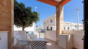 Un balcon avec deux chaises en plastique blanc et une table, un petit meuble blanc et un arbre qui offre de l’ombre.