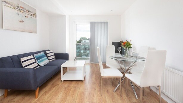A modern living room with a dark blue sofa, white chairs, a glass coffee table, and a dining table with white chairs. A painting is on the wall, and a window with curtains is visible. - Skyvillion Apartments - TowerPoint Enfield - 1Bed Deluxe Apartment w/ Balcony (Greater London)