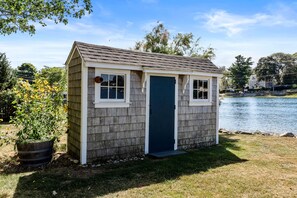 A small, single-story shed with a dark blue door and white trim, situated on a grassy area near a body of water.