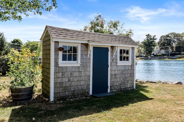 A small, single-story shed with a dark blue door and white trim, situated on a grassy area near a body of water.