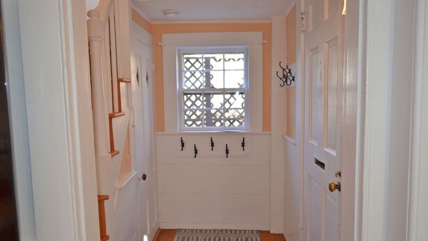 Un pasillo estrecho con puertas blancas, una ventana con celosía y un felpudo estampado.