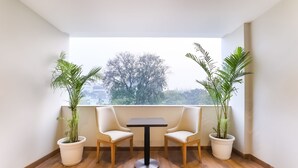 Un balcón con dos sillas y una mesa, rodeado de plantas en macetas, con vistas a árboles y edificios.