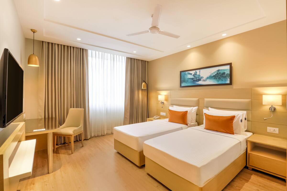 A hotel room with two beds, a desk, a chair, a TV, and a ceiling fan.