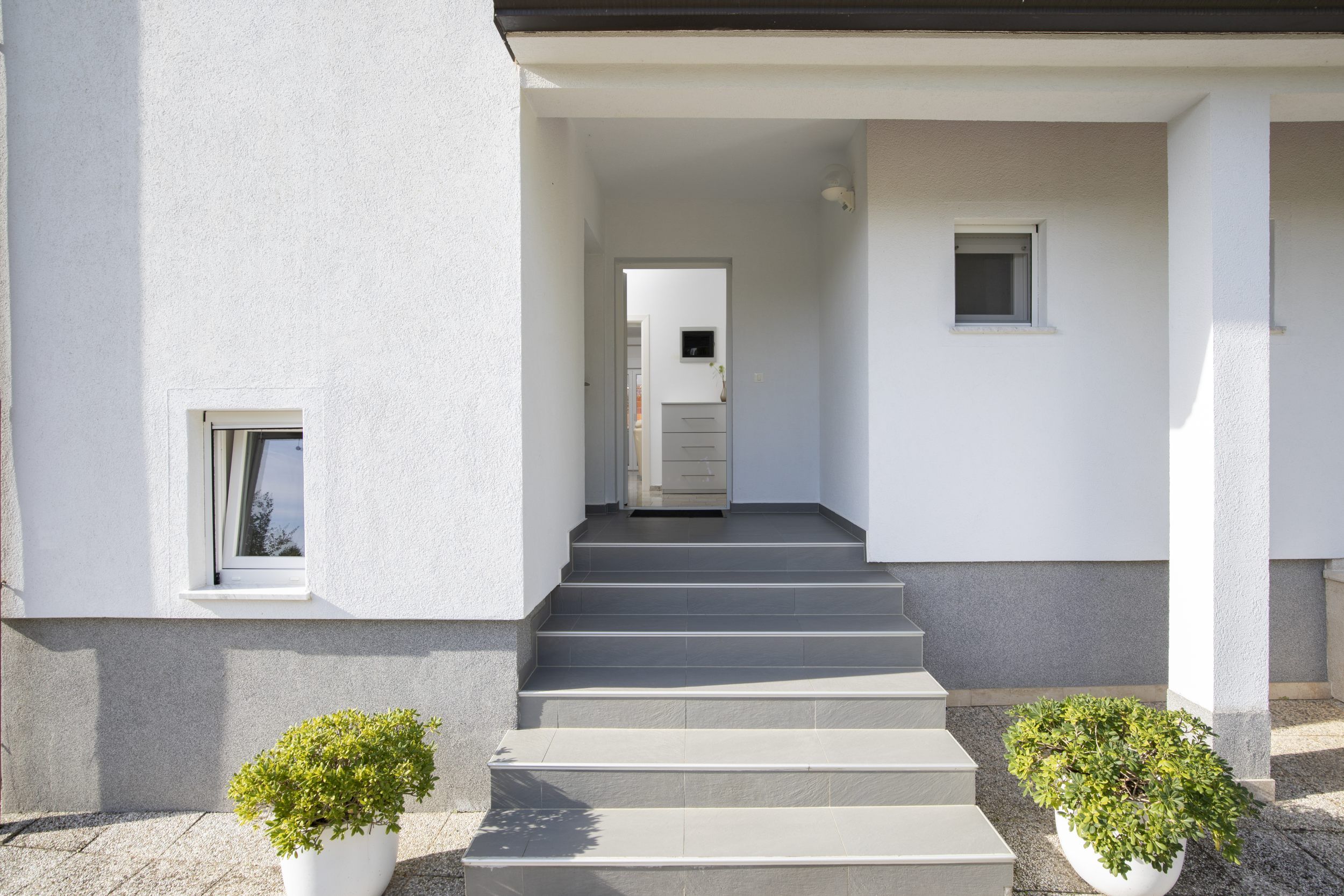 Un bâtiment moderne avec un escalier en béton menant à une porte. De part et d’autre des marches, on trouve deux plantes en pot.