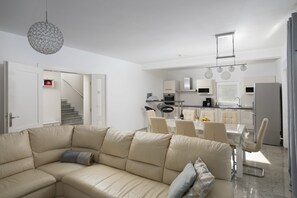 A modern living room with a beige sectional sofa, a dining area with a table and chairs, a kitchen with white cabinets, and a staircase leading to another level.