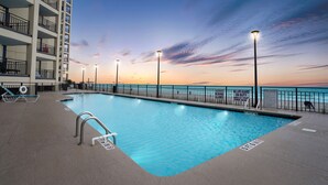 Piscina com vista para o oceano ao pôr do sol, cercada por uma grade e postes de iluminação.