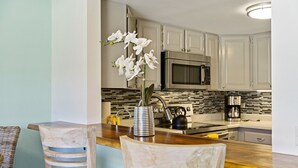 A kitchen with white cabinets, a microwave, and a stainless steel stove. There is a wooden bar counter with a vase of white flowers. - Summer Deal Beachfront 2BR Kona Condo Across from Magic Sands Beach (Kailua Kona)