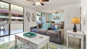 A living room with a grey sofa, a white coffee table, a white side table, and a ceiling fan. - Summer Deal Beachfront 2BR Kona Condo Across from Magic Sands Beach (Kailua Kona)