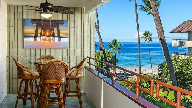A balcony with wicker furniture, a ceiling fan, and a painting of a sunset over water. - Summer Deal Beachfront 2BR Kona Condo Across from Magic Sands Beach (Kailua Kona)
