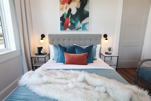 Une chambre à coucher avec une tête de lit capitonnée, un grand tableau abstrait, deux tables de chevet avec des lampes et une couverture en fausse fourrure blanche.