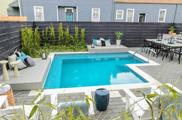 Une piscine rectangulaire, bordée d’une terrasse carrelée, entourée d’une clôture sombre, et un espace patio aménagé avec des meubles d’extérieur et des plantes.