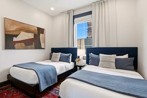 A hotel room with two beds, a large abstract painting, a window with curtains, and a nightstand with a lamp and flowers. - 7BR City Condo | Pool Access Near CBD (New Orleans)
