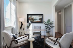 Una sala de estar moderna con una mesa de vidrio, dos sillones, un tapete negro, un cuadro enmarco, una lámpara y una planta en maceta.