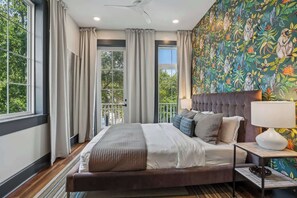 A bedroom with a large bed, a nightstand, a lamp, and a view of greenery through the window. - 7BR City Condo | Pool Access Near CBD (New Orleans)