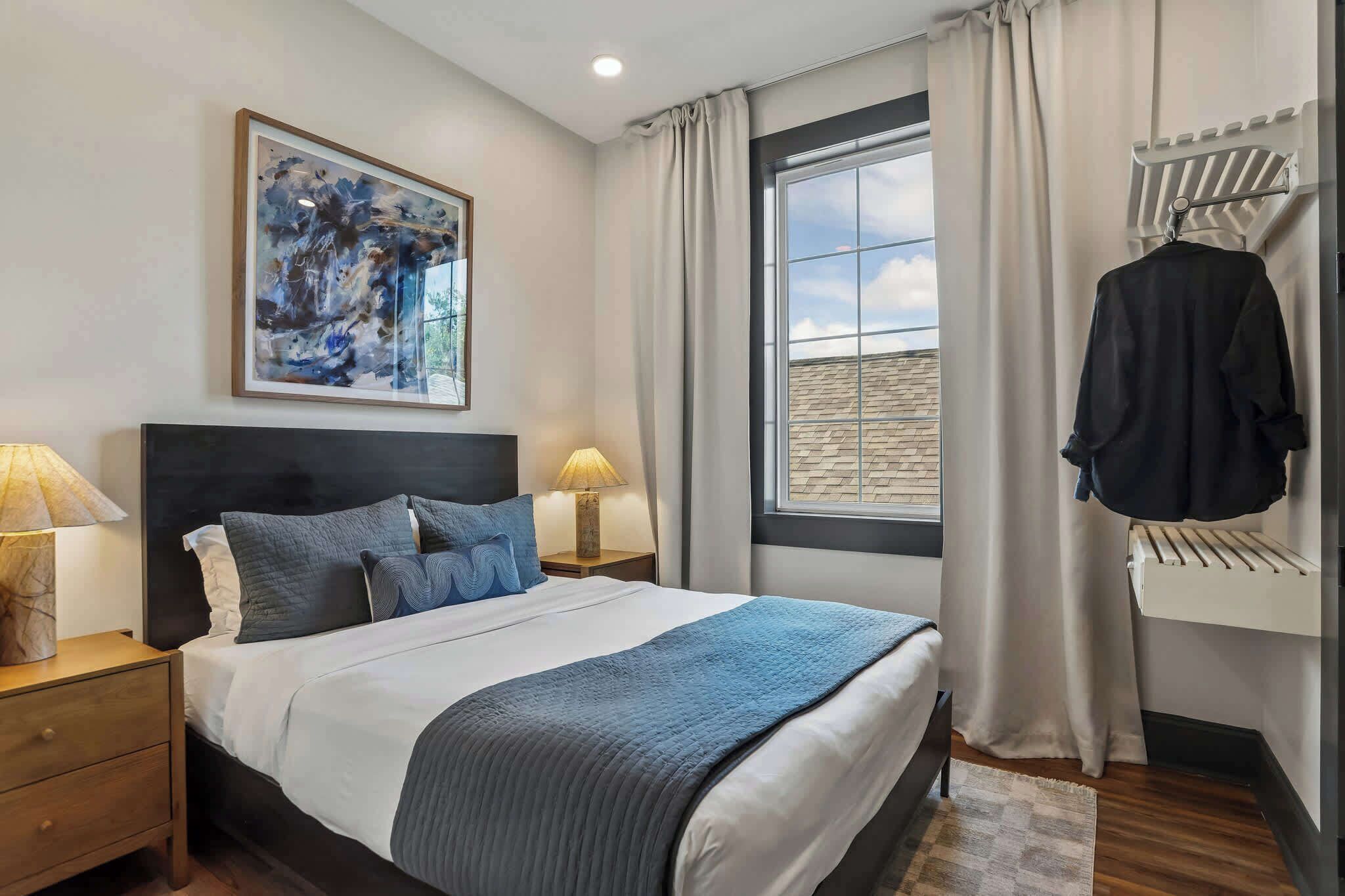 Un dormitorio con cama, dos mesitas de noche, una ventana con cortinas, un perchero y un cuadro en la pared.