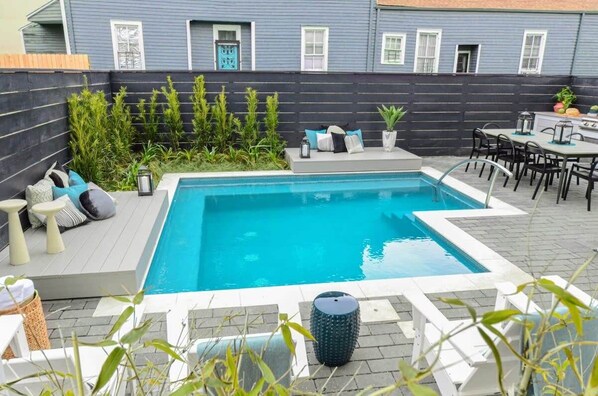 Una piscina rectangular con solárium empedrado, rodeada por una cerca oscura, y un patio con mobiliario exterior y plantas.