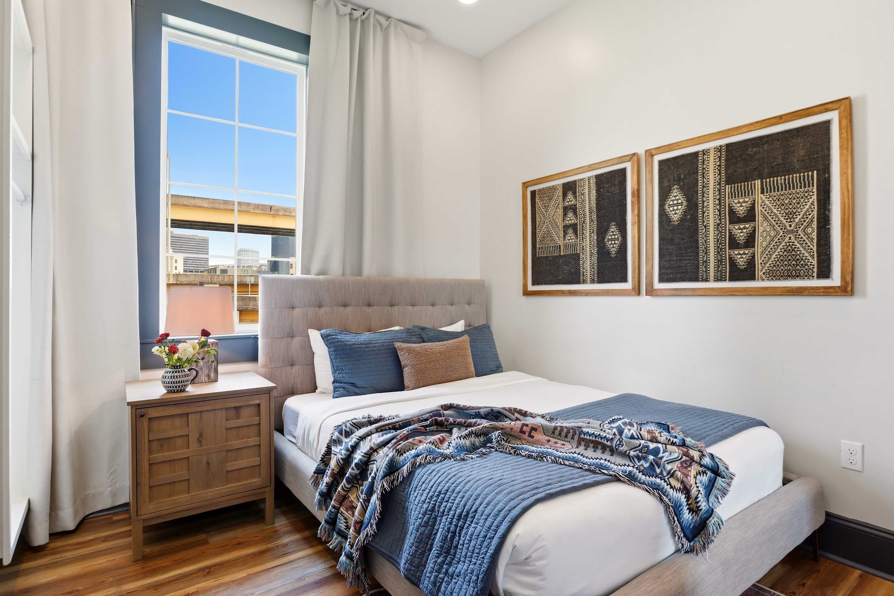 A bedroom with a bed, a nightstand, a window with curtains, and two wall-mounted artworks.