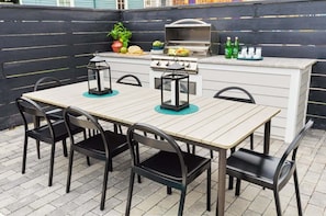 Un patio con mesa de madera y sillas negras, una parrilla y una pequeña zona de bar con botellas y vasos.