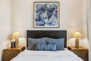 A neatly made bed with a dark headboard, flanked by two bedside tables with lamps, and a framed abstract painting above.