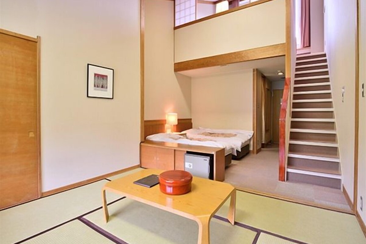A hotel room with a bed, a desk, a chair, a small table, a picture on the wall, and a staircase leading to an upper floor.