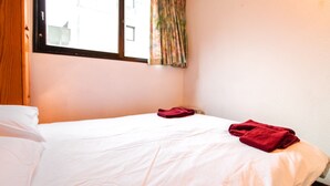 Uma cama de solteiro com roupa de cama branca e toalhas vermelhas, uma janela com cortinas e uma porta de madeira.