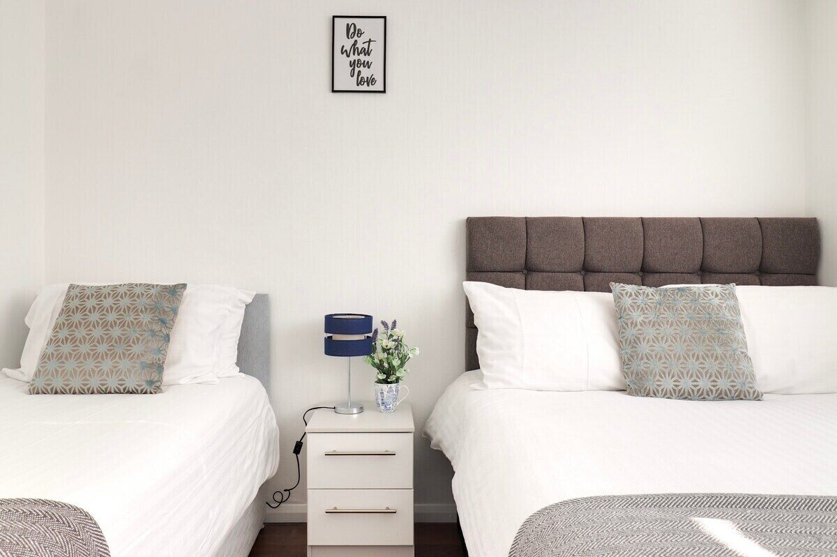 Quarto de hotel com duas camas, mesa de cabeceira com luminária e um vaso de flores, e uma frase motivacional na parede.
