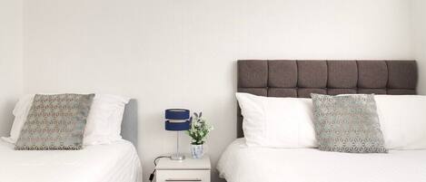 Quarto de hotel com duas camas, mesa de cabeceira com luminária e um vaso de flores, e uma frase motivacional na parede.