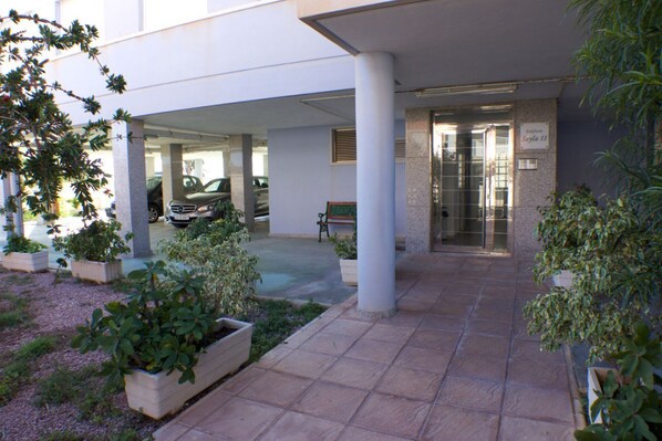 A covered outdoor area with a tiled floor, potted plants, a bench, and a car parked under a building.