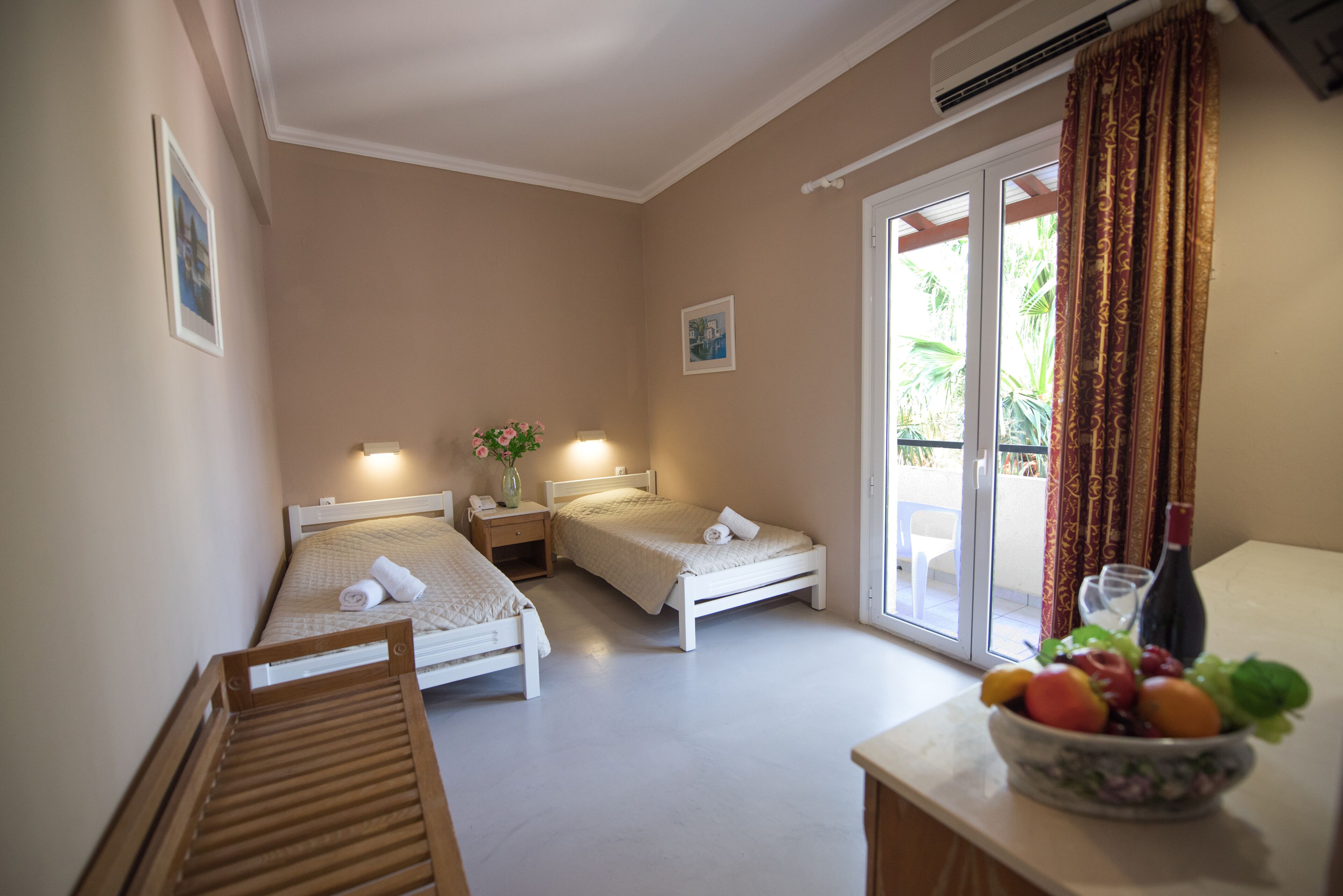 A small, well-lit bedroom with two beds, a wooden chair, a nightstand, a vase with flowers, and a fruit bowl on a table.