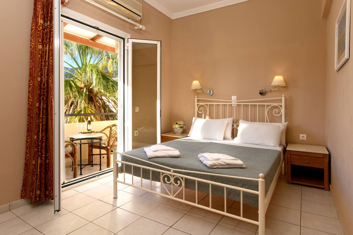 A bedroom with a bed, two bedside lamps, a nightstand, and a view of a balcony with a palm tree.