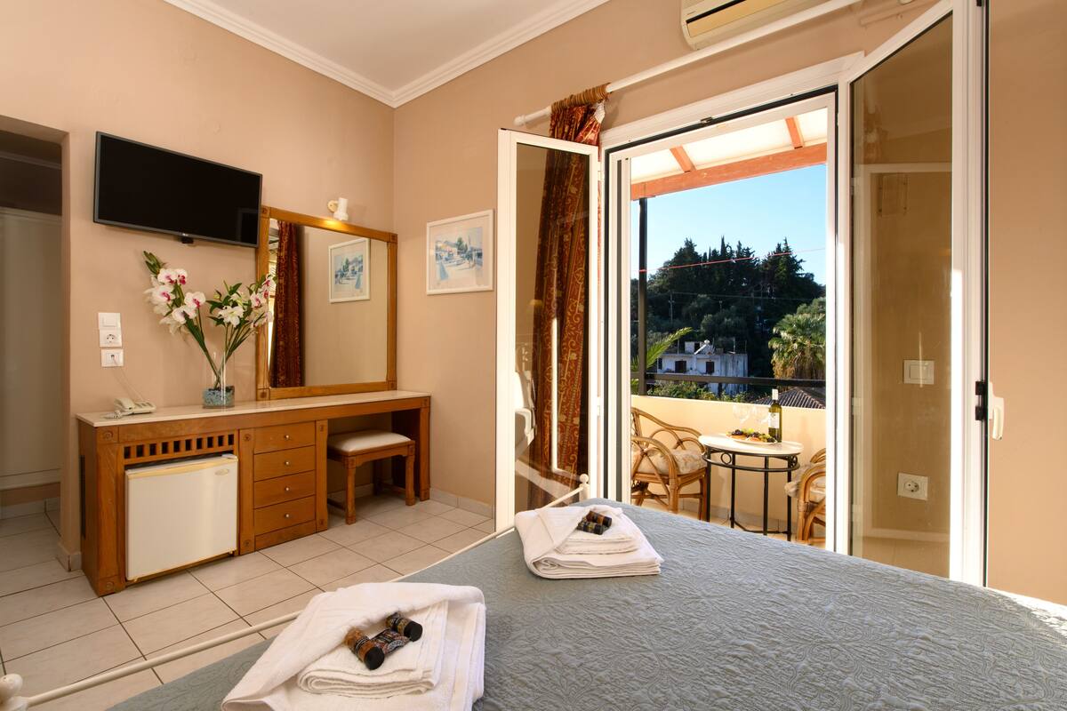 A hotel room with a bed, a desk, a mirror, a TV, and a balcony with a table and chairs.