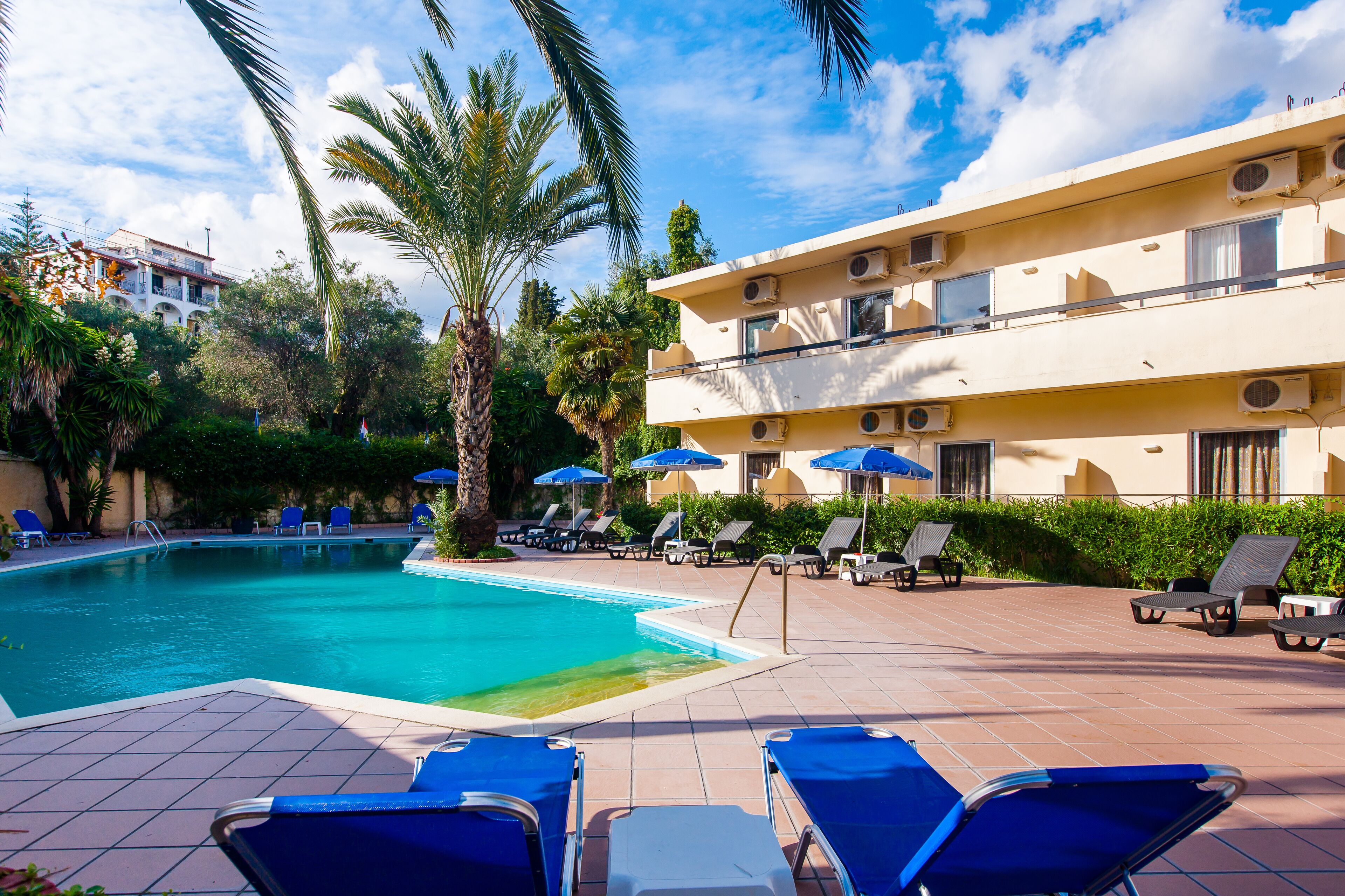 Una piscina con sillas de descanso y sombrillas, rodeada de palmeras y un edificio con balcones.
