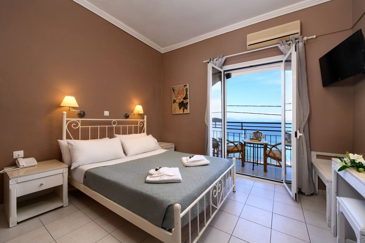 A hotel room with a bed, bedside lamps, a desk, a chair, and a balcony with a view of the sea.
