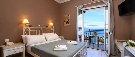 A hotel room with a bed, bedside lamps, a desk, a chair, and a balcony with a view of the sea.