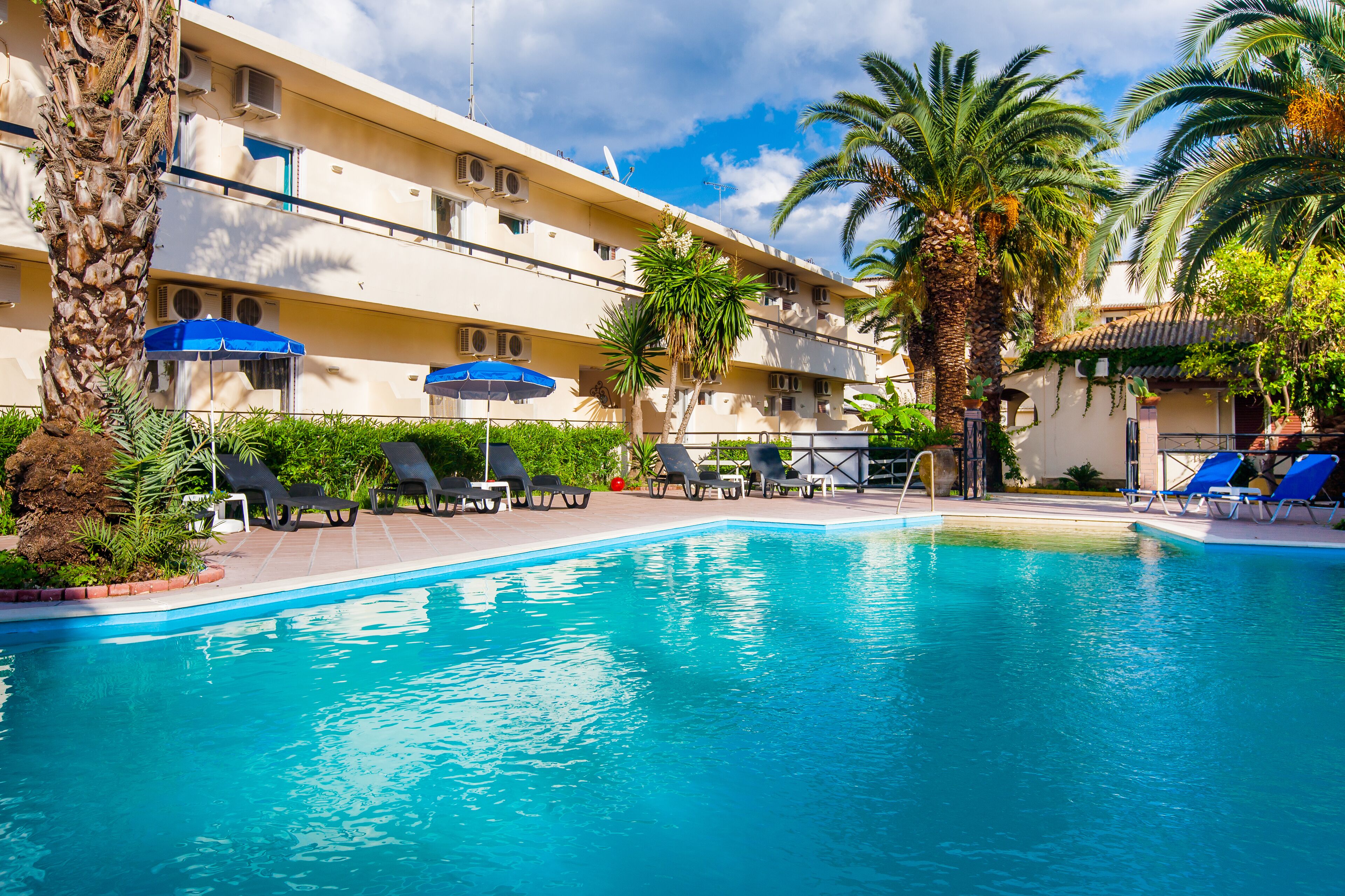 Una piscina con sillas de descanso y sombrillas, rodeada de palmeras y un edificio con varios balcones.