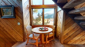 Acogedora habitación tipo cabaña de madera con una mesa redonda y dos sillas, un cuadro en la pared y una ventana con vistas a árboles y montañas.