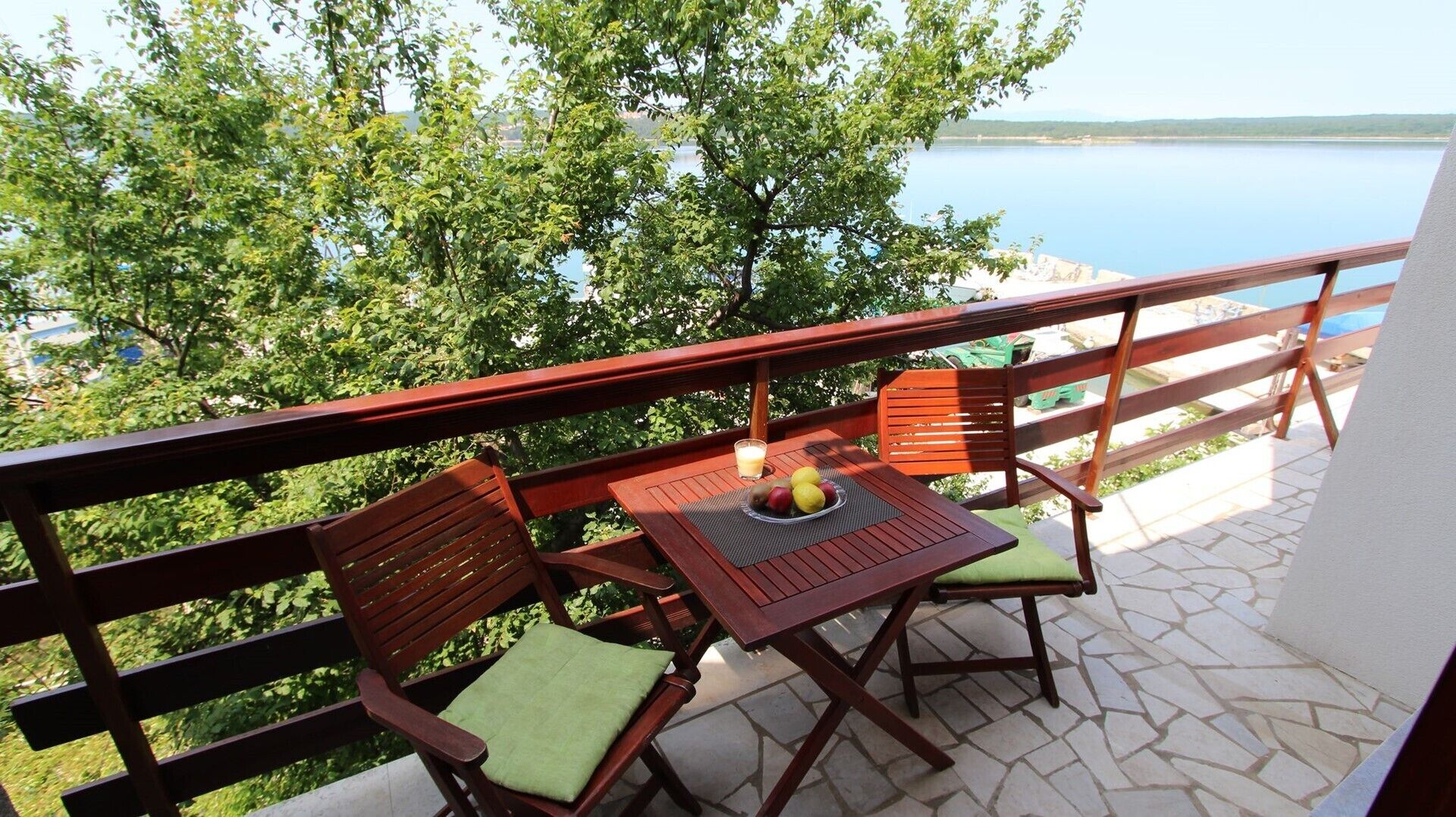 Un balcone con due sedie di legno e un tavolo, un bicchiere di succo e una ciotola di frutta.
