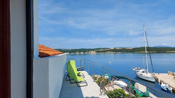 Ein Balkon mit Blick auf einen stillen See, ein vor Anker liegender Segelboot und ein grüner Stuhl.