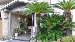 A building entrance with a stone facade, a ramp leading up, and large tropical plants. - Hinode Ryokan (Fukui)