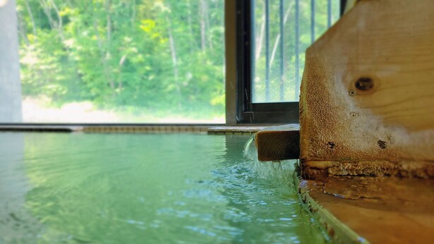 A wooden edge of a pool with water flowing into it, viewed from inside a building with a window overlooking a forest. - Shikanoyu Tsuchiya (Tsumagoi)